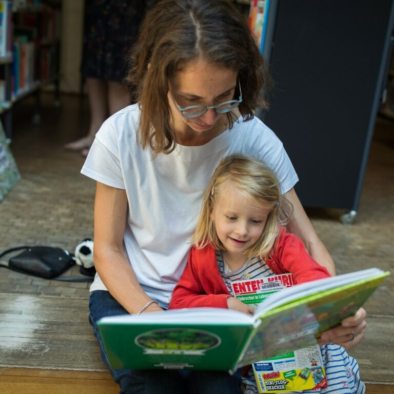 Eine Frau liest mit einem Kleinkind auf dem Schoß ein Buch in einer Bibliothek.