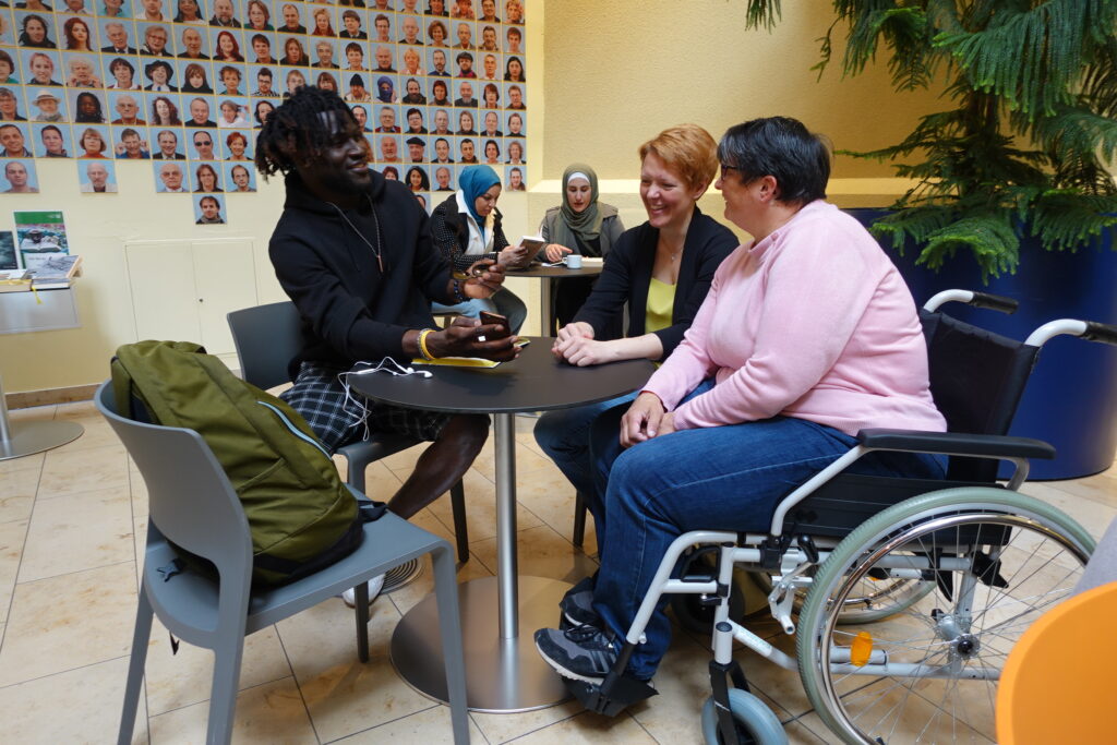 Britta Schmedemann sitzt an einem Tisch in einem Raum. Links von ihr sitzt ein Mann mit schwarzer Hautfarbe, rechts eine Frau mit weißer Hautfarbe und im Rollstuhl. Im Hintergrund sind weitere Personen zu sehen.