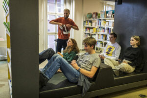 Vier Jugendliche sitzen in einer Bibliothek und spielen mit einer Spielkonsole. Hinter ihnen ein Bibliothekar und Bücherregale.
